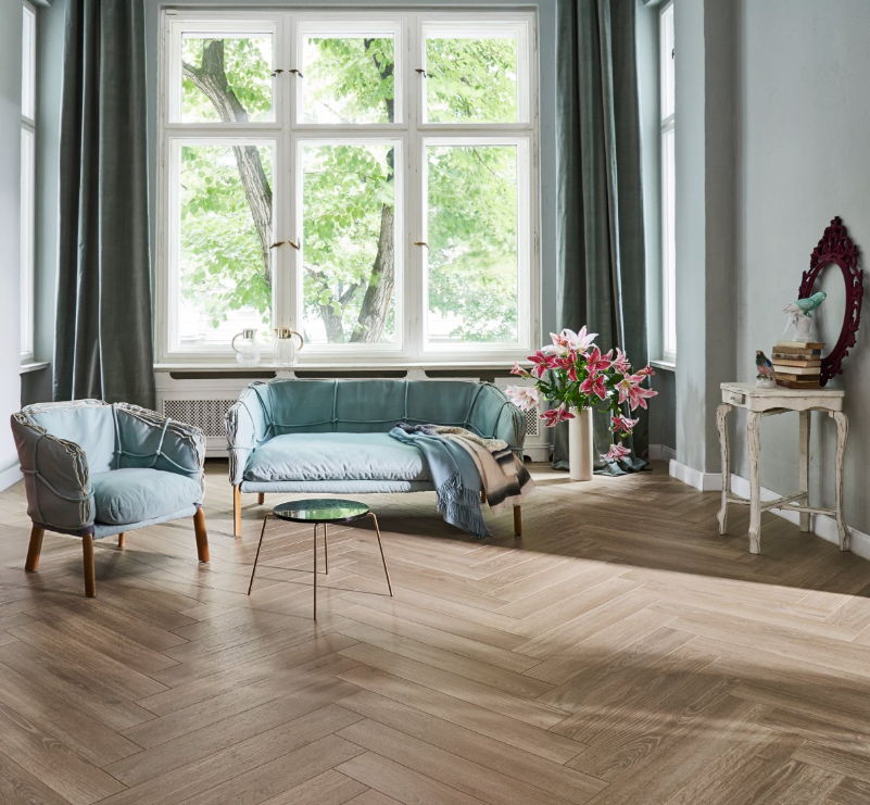 Living room with a blue sofa, armchair, and coffee table in front of a large window featuring herringbone wooden flooring
