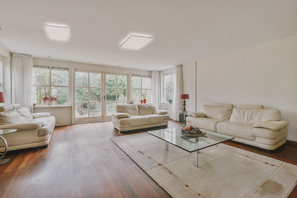 Two LED infrared heaters on the ceiling of a modern living room with beige sofas, a glass coffee table, and large windows.